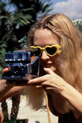 Blonde woman in heart-shaped sunglasses taking photo with vintage Polaroid camera outdoors.