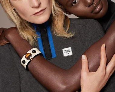 Two models embracing, wearing geometric jewelry and a knit top with a label that says "Opening Ceremony."