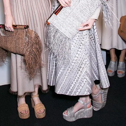 Fashionable individuals in textured, neutral dresses holding woven and fringed clutches, wearing platform sandals.