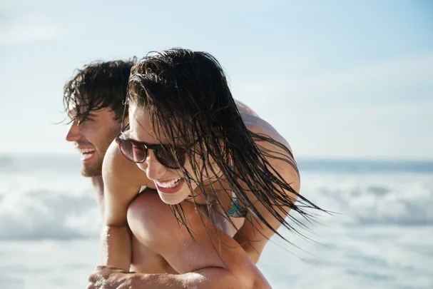 A couple laughing and enjoying a piggyback ride on a sunny beach.