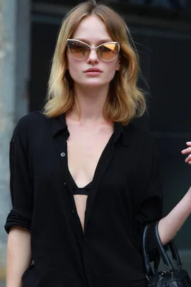 Woman with blond hair wearing cat-eye sunglasses, a black open shirt, and carrying a handbag.