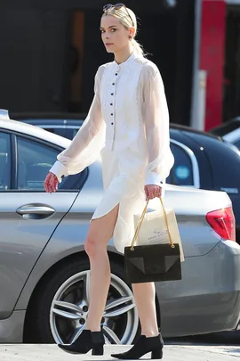 A person in a white dress and black boots walks on a street, holding a green handbag and a shopping bag.