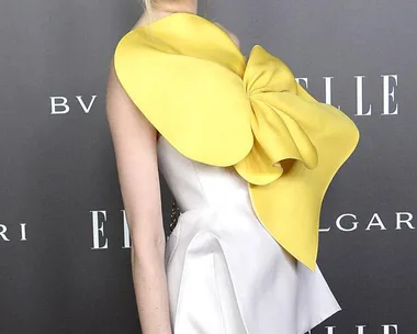 A woman in a white top with a large yellow bow stands on the red carpet at an event, with ELLE and Bulgari logos behind her.