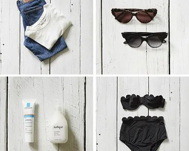 Folded jeans and t-shirt, two pairs of sunglasses, skincare bottles, and a black bikini on white wood.