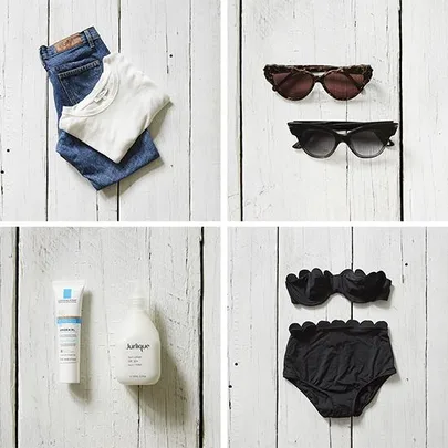 Folded jeans and t-shirt, two pairs of sunglasses, skincare bottles, and a black bikini on white wood.