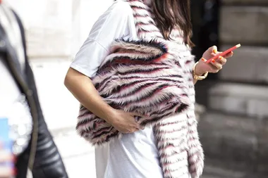 A person wearing a white shirt and a striped faux fur vest holds a red smartphone.