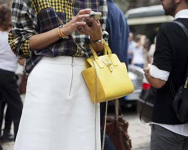 Person in plaid shirt and white skirt holding a phone, wearing a yellow handbag, in a crowded street.