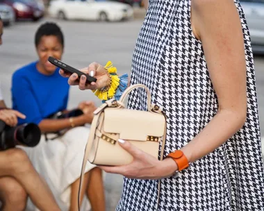 Woman in houndstooth dress holds phone and handbag, two people sit blurred in background on street.