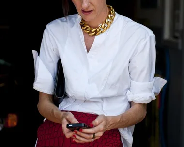 A woman wears a white blouse, bold gold necklace, and red skirt, holding a smartphone and looking down.