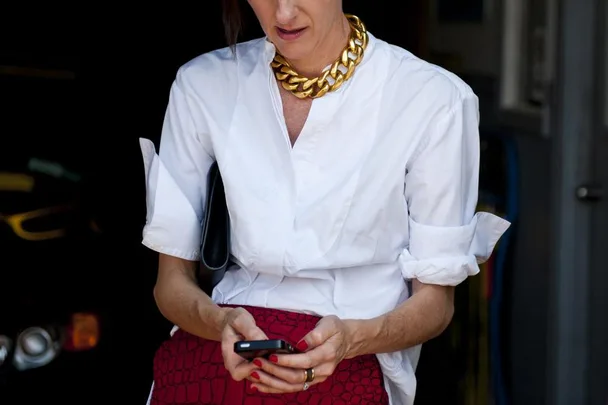 A woman wears a white blouse, bold gold necklace, and red skirt, holding a smartphone and looking down.