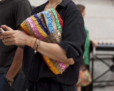 Person holding a phone and a colorful striped clutch handbag, wearing a black outfit with bracelets and an orange string on the wrist.