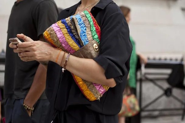 Person holding a phone and a colorful striped clutch handbag, wearing a black outfit with bracelets and an orange string on the wrist.