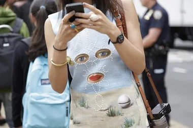 A woman wearing a dress with a surreal face design uses a smartphone on a city street, with a police officer in the background.