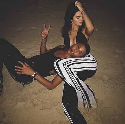 Two people sitting on the sand at night, one flashes a peace sign, both in stylish outfits.