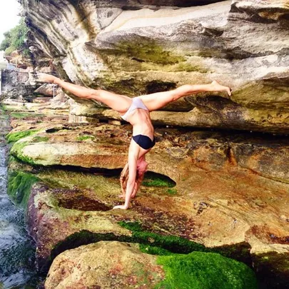 Person doing a handstand on green rocks beside a cliff, wearing a black and gray bikini.