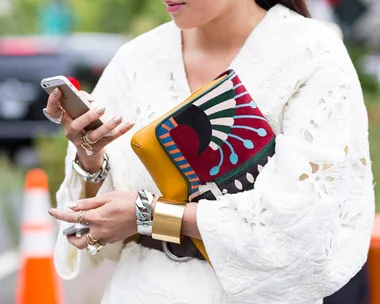 A person in a white dress holding a phone and a colorful clutch bag with a distinct geometric pattern.