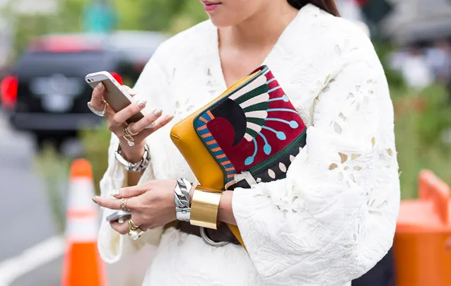 A person in a white dress holding a phone and a colorful clutch bag with a distinct geometric pattern.