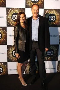 Two people, a woman in a black dress and leather jacket and a man in a suit, pose together at a formal event.