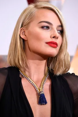 A woman with blond hair and red lipstick wearing an elegant blue and gold necklace against a black outfit at an event.