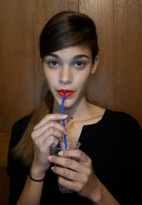 A person with red lipstick sipping a drink through a blue straw from a clear cup.