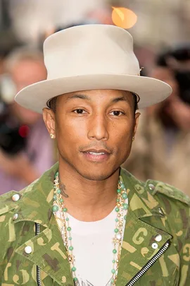 Pharrell Williams wearing a green patterned jacket, white hat, and layered necklaces while smiling.