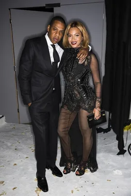 A man in a suit and a woman in a black dress pose together, smiling, at a fashion event.