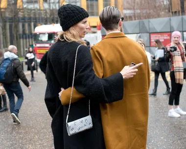 A couple in coats stand close together outdoors, with a bokeh background of people, buildings, and a truck.