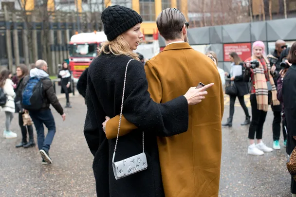 A couple in coats stand close together outdoors, with a bokeh background of people, buildings, and a truck.