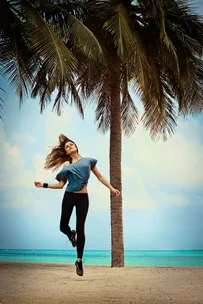 Person in workout gear joyfully jumping near a palm tree on a beach with turquoise water and a blue sky in the background.