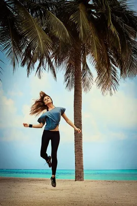 Person in workout gear joyfully jumping near a palm tree on a beach with turquoise water and a blue sky in the background.