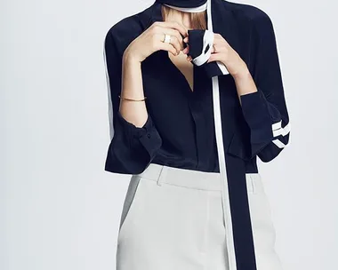 A woman in a black blouse with white trim and white trousers poses against a plain background.