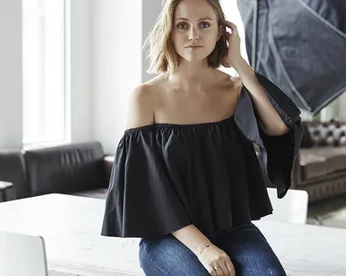 Woman in black off-the-shoulder top and jeans, sitting on white table in studio with large softbox light behind her.