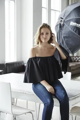 Woman in black off-the-shoulder top and jeans, sitting on white table in studio with large softbox light behind her.