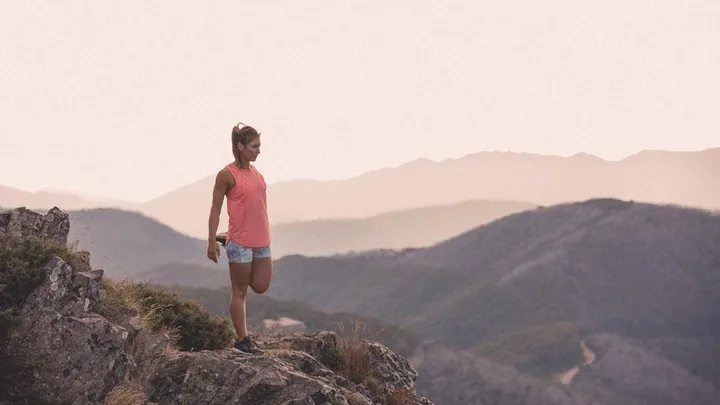 Woman in athletic wear stretching on a rocky hilltop with distant mountains in the background.