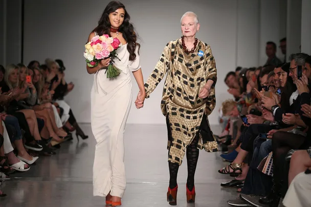 Vivienne Westwood and her granddaughter Cora Corre walking down a runway holding hands. Cora holds a bouquet of flowers.