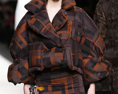 Model on runway wearing oversized padded jacket, mini skirt with geometric brown and black pattern, carrying Fendi pompom accessory.