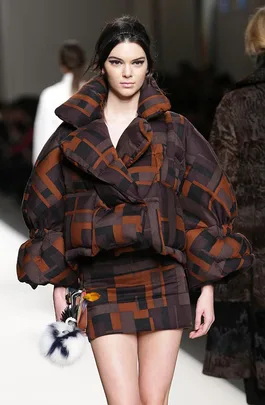 Model on runway wearing oversized padded jacket, mini skirt with geometric brown and black pattern, carrying Fendi pompom accessory.