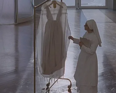 A woman dressed as a nun looks at a white dress hanging on a rack in a sunlit room.