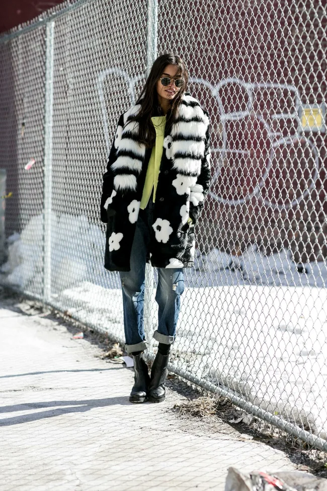 Woman in a patterned coat, yellow top, jeans, and boots stands by a chain-link fence in a snowy urban setting.