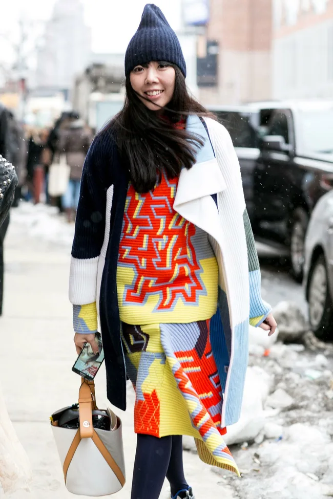 A woman in a colorful knitted outfit and a beanie, holding a bag and phone, walks outside on a snowy day.