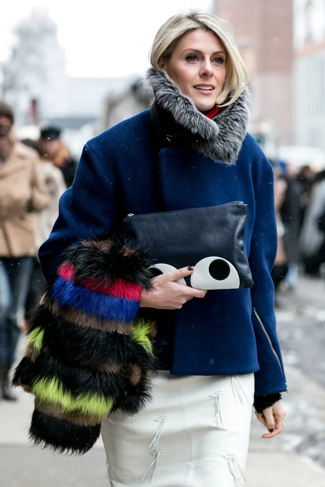 Person in a blue coat with gray fur collar, holding a quirky clutch, wearing a colorful fur item, walking on a snowy street.