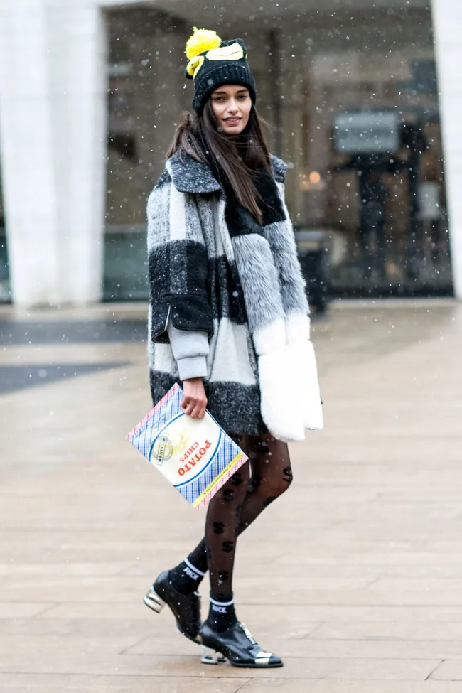 Young woman wearing a stylish coat and beanie, holding a bag of potato chips in light snowfall.