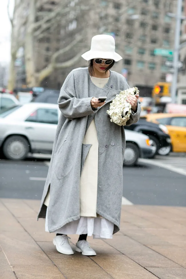 A person in a grey coat and white hat, looking at a phone, on a city street with taxis in the background.