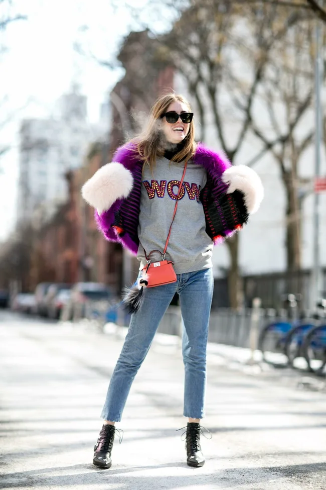 A woman smiles in a gray "WOW" sweater, purple fur coat, sunglasses, jeans, and boots on a sunny street.