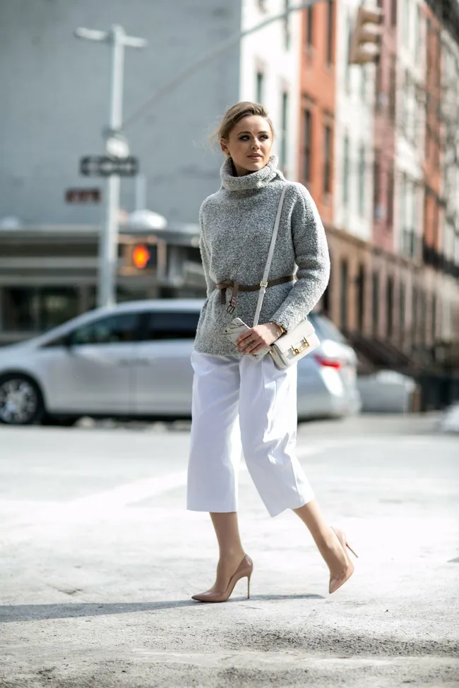 Stylish woman in a gray sweater and white pants walking on a city street in heels, holding a white handbag.