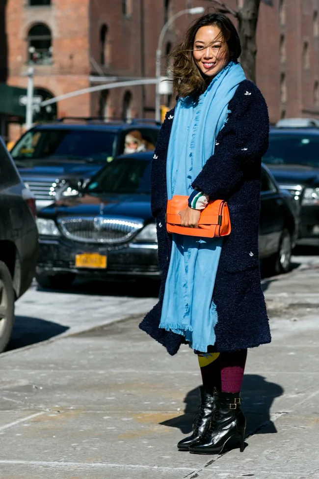 Woman in a navy coat and blue scarf smiles on a city street, holding an orange clutch.