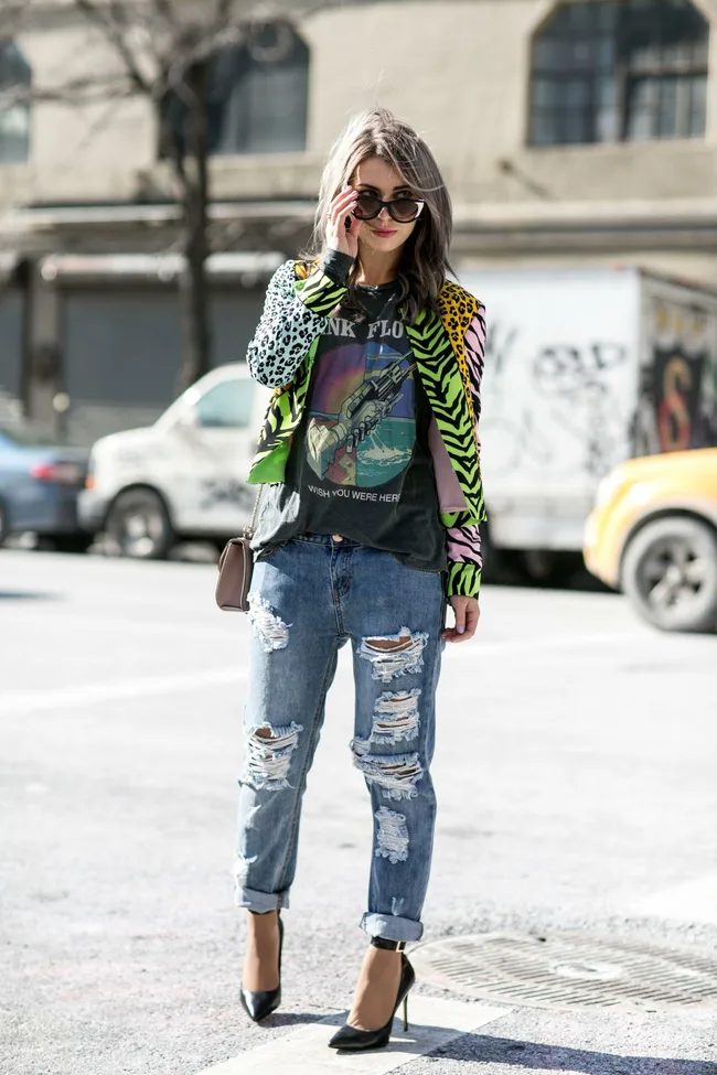 Woman with gray hair in sunglasses wearing a Pink Floyd shirt, colorful jacket, ripped jeans, and black heels.
