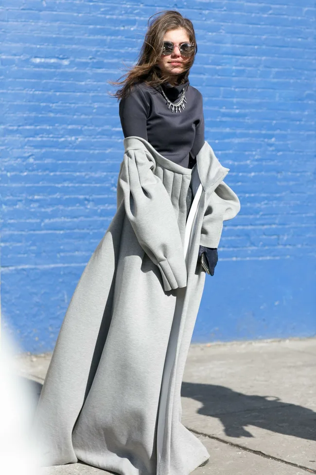 Woman in sunglasses wearing a gray coat over a black top, standing outdoors against a blue brick wall.