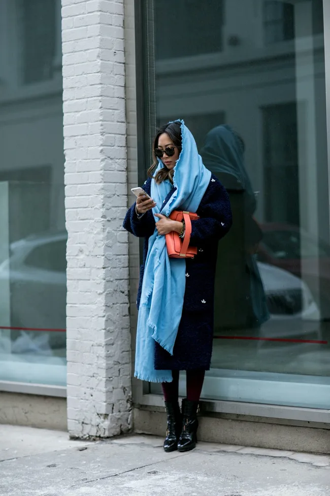 Person in a blue coat and scarf stands by a window, holding a phone and orange clutch.