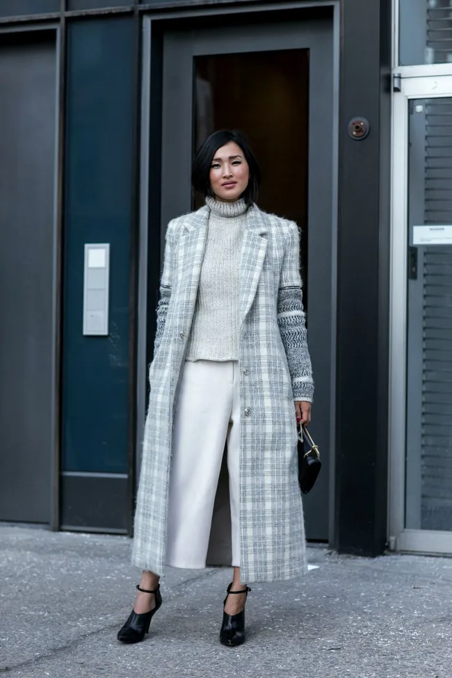 Woman in a plaid coat and knit sweater stands outside a building, holding a small black handbag.
